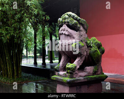 Bemoosten Stein Lion vor einem chinesischen Gebäude mit traditioneller Architektur, neben Bambus Bäume. Stockfoto