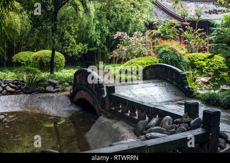 Steinerne Brücke in einem Chinesischen Garten. Traditionelle Gartengestaltung mit Stein Brücke über einen kleinen Teich, der von Bäumen und Blumen an einem regnerischen Tag umgeben. Stockfoto