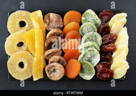 Kandierte Fruchtstücke von Papaya, Kiwi, Ananas, getrocknete Feigen, Datteln, Aprikosen Stücke kandierten Ingwer Wurzel, der lag auf einem schwarzen Keramikplatte auf einem schwarzen Stockfoto