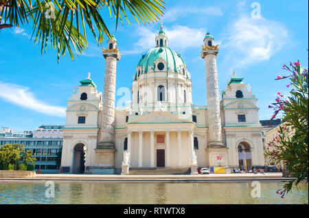 Fassade der Karlskirche (St. Charles Kirche) gegen den blauen Himmel, weiße Wolken und ein Teich mit Wasser an einem heißen Sommertag. Grün Zweig der Baumstruktur mit rosa Flo Stockfoto