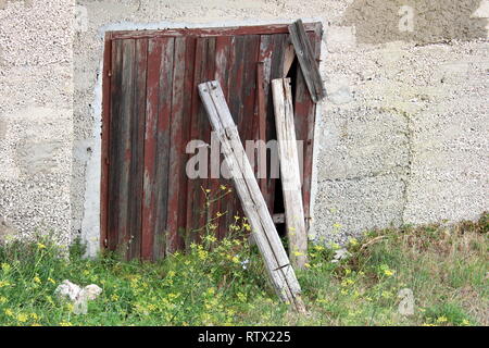Verlassenes Haus Wand mit alten gerissenen Holztür Garagentore mit Risse im Lack überzogen und mit Holzbrettern mit ungeschnittenem Gras umgeben unterstützt. Stockfoto