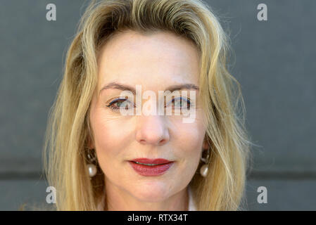 Attraktive blue eyed blonde Frau trug Make-up in einer Nahaufnahme Kopf geschossen gegen einen grauen Außenwand Stockfoto