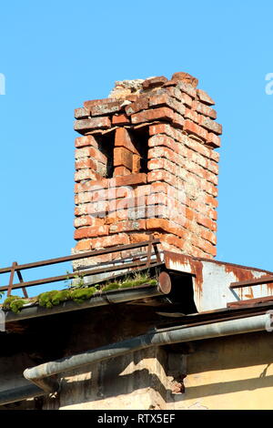 Die Ruinen der alten roten gemauerten Schornstein auf verlassenen Gebäude mit Metalldach und verrostet Kante mit dachrinne am klaren, blauen Himmel Hintergrund umgeben Stockfoto