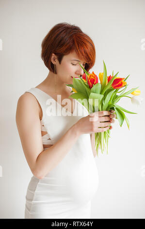 Glückliche junge schwangere Frau in weißem Kleid mit Blumen Stockfoto