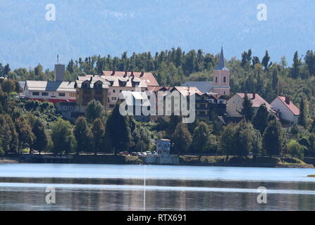 Kleine Bergstadt mit hohen dicht bebaute Familie Häuser rund um die Kirche mit klaren See vor und dichte Bäume mit Berg im Hintergrund Stockfoto