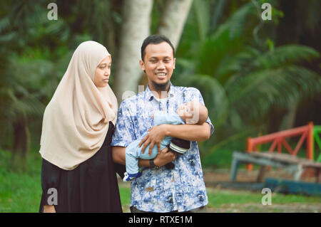 Asiatische muslimischen hijabi Mutter und Vater zu Fuß durch den Park mit Sohn im Kinderwagen, während seine Mutter kümmert sich um Ihr Baby Stockfoto