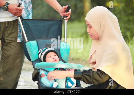 Asiatische muslimischen hijabi Mutter und Vater zu Fuß durch den Park mit Sohn im Kinderwagen