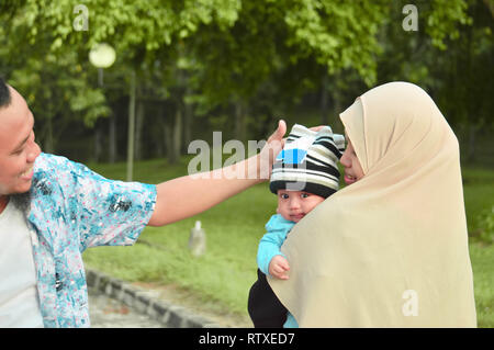 Asiatische muslimischen hijabi Mutter und Vater zu Fuß durch den Park mit Sohn im Kinderwagen, während seine Mutter kümmert sich um Ihr Baby Stockfoto