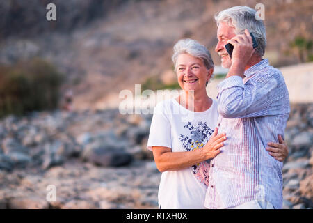 Konzept der Ferienhäuser, Technologie, Tourismus, Reisen und Menschen - gerne älteres Paar mit Mobiltelefon am Telefon auf Kiesel-strand Lachen und Scherzen. Wh Stockfoto