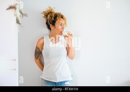 Attraktive lockige Blondine mittleren Alters niedliche Frau trinkt Wasser und Zitrone zu Hause mit weisse Wand im Hintergrund - gesunder Lebensstil für cacuasian Menschen Stockfoto