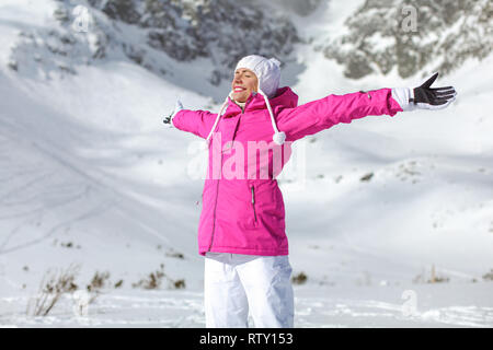 Junge Frau in Rosa ski Jacke, Handschuhe und Hosen, die Arme ausgebreitet, die Augen geschlossen, lächelnd vor Sonne, Schnee bedeckten Berg hinter ihr. Stockfoto