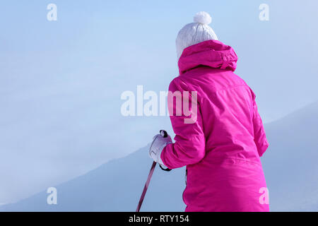 Von hinten betrachten, junge Frau in rosa Winterjacke, Stöcke in die Hände, um den Hügel hinunter zu gehen. Stockfoto