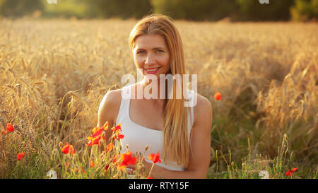 Junge Frau sitzt im Weizenfeld, beleuchtet durch die Nachmittagssonne, Paar roter Mohn um Sie im Vordergrund. Stockfoto