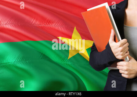 Lernen Sprache Konzept. Junge Frau mit den Burkina Faso Flagge im Hintergrund. Lehrer, orange leer Buchen Sie abdecken. Stockfoto