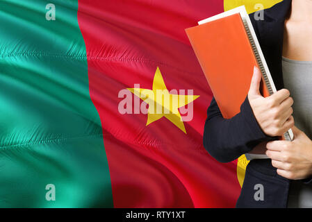 Lernen kamerunischen Sprache Konzept. Junge Frau mit den Kamerun Flagge im Hintergrund. Lehrer, orange leer Buchen Sie abdecken. Stockfoto