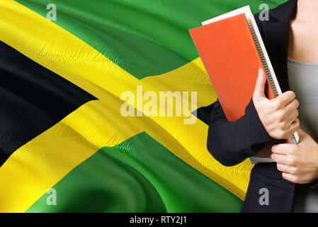 Lernen jamaikanische Sprache Konzept. Junge Frau mit den Jamaika Flagge im Hintergrund. Lehrer, orange leer Buchen Sie abdecken. Stockfoto