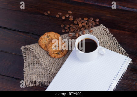 Weiße Tasse Kaffee mit Kaffee Körner in der Nähe und Keksen und Notizblock in einem Käfig für Aufzeichnungen auf Leinen Stoff auf der dunklen vintage Hintergrund. Stockfoto