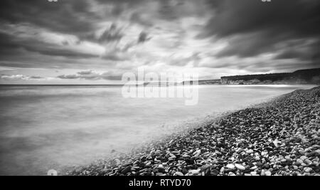 Schwarz-weiß Bild von einer stürmischen Himmel und welliges Meer mit Wellen schlagen der am Ufer des Meeres voll mit schönen Steinen. Longexposure Fotografie Stockfoto