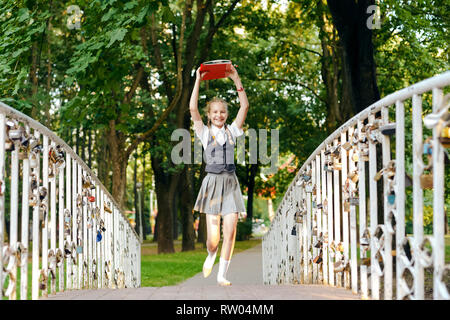 Student Schülerin mit Zöpfen in Uniform mit Bücher in die Hände über dem Kopf läuft glücklich über die Brücke im Sommer Park Stockfoto