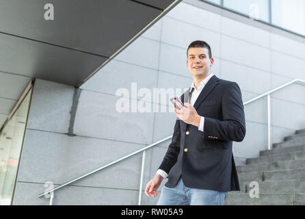 Junge und Casual Geschäftsmann Sie Ihr Smartphone während des Gehens außerhalb des Bürogebäudes Stockfoto