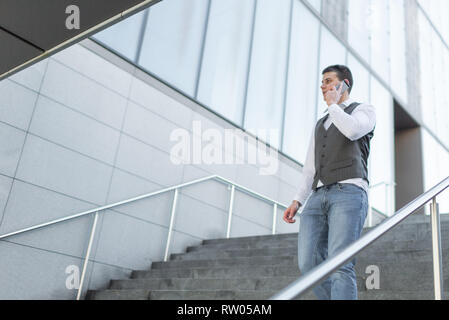 Junge und Casual Geschäftsmann Sie Ihr Smartphone während des Gehens außerhalb des Bürogebäudes Stockfoto