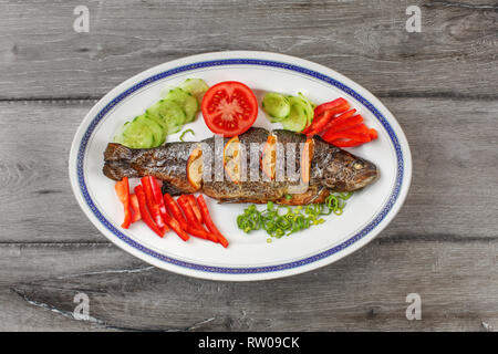 Tischplatte, gegrillte Forelle auf dem ovalen Schild, Orangenscheiben, Tomaten, Paprika, Gurken und Frühlingszwiebeln. Stockfoto