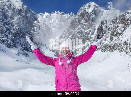 Junge Frau in Rosa ski Jacke, Handschuhe und Winter hat, lächelnd, werfen Schnee in der Luft, Sonne auf den Berg hinter ihr leuchtendes. Skalnate Pleso Skifahren re Stockfoto