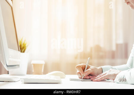Eine Frau schreibt ein Stift auf einem Blatt Papier. Am Computer arbeiten, Kaffee auf dem Tisch im Büro zu Hause, close-up. Sonnenlicht im Fenster Stockfoto