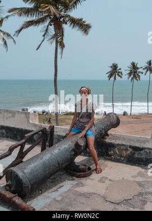 Ein Foto von einem Europäischen Frau auf eine riesige Kanone auf Elmina Castle posieren. Ozean können auf dem Hintergrund gesehen werden. Stockfoto