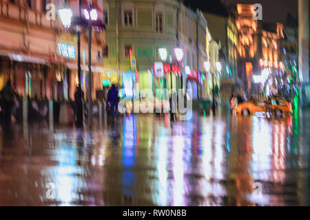 Beeilen, um jungen Menschen unter Sonnenschirmen an einem regnerischen Nacht in der Straße der Stadt. Brillante Ausleuchtung von Laternen und Schaufenstern. Konzept der Jahreszeiten Stockfoto