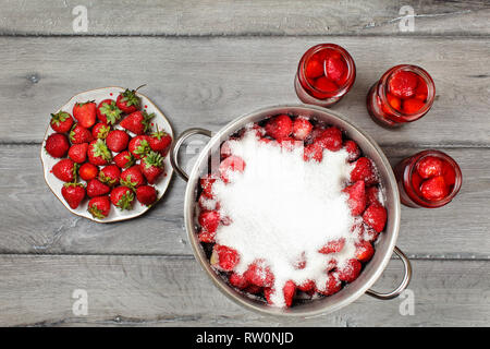 Tischplatte, groß, Stahl Topf mit Erdbeeren in Kristallzucker abgedeckt, drei Flaschen mit Früchten in Sirup und Platte. Hausgemachte Erdbeere Stockfoto