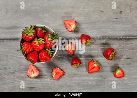 Tischplatte - kleine Keramik Schale Erdbeeren, mehr geschnittenes Obst herum liegen auf grau Holzschreibtisch. Stockfoto