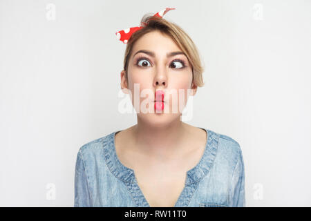 Closeup Portrait von Lustig verrückt junge Frau in lässigem Blue Denim Shirt mit Make-up und roten Stirnband stehend, gekreuzte Augen mit Fisch Lippen Geste. ind Stockfoto