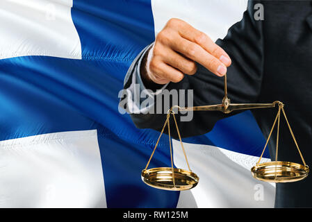 Finnische Richter goldene Waage der Gerechtigkeit mit Finnland wehende Flagge Hintergrund. Gleichstellung von Frauen und Männern Thema und rechtlicher Begriff. Stockfoto