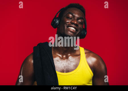 Afrikanischer Mann mit Handtuch über der Schulter genießen Hören von Musik über Kopfhörer. Passen junge Mann Musik hören während des Trainings Pause auf roten Hintergrund. Stockfoto