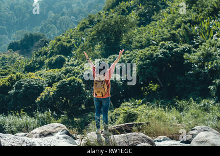 Asiatische Frau Reisenden stehen auf Rock und die Arme in die Luft im Querformat der grünen Regenwald und Strom Wasser im sonnigen Tag im Wald. Backpacker Stockfoto