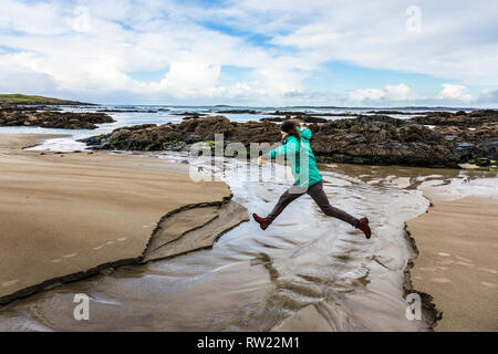 Ardara, County Donegal, Irland. 4. März 2019. Eine Frau springt über einen Bach an einem Strand auf den 'wilden Atlantischen Weise". Der Tag ist kalt und windig. Credit: Richard Wayman/Alamy leben Nachrichten Stockfoto