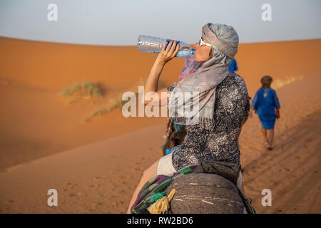 Eine Frau, die Getränke aus einer grossen Flasche Wasser, während auf einem Kamel reiten, Merzouga, Marokko Sahara-Erg Chabbi Dünen Stockfoto