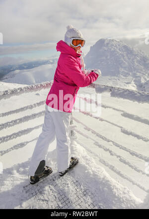 Junge Frau in Rosa ski Jacke, Hosen, Schuhe, Mütze und Handschuhe, lehnte sich auf Schnee bedeckten Rampe, lächelnd. Stockfoto