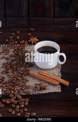 Weiße Tasse Kaffee mit Kaffee Körner in der Nähe von auf Leinen Stoff auf der dunklen vintage Hintergrund. Vertikale Foto. Stockfoto