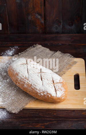 Frisches Brot auf einer Eiche board mit verstreuten Mehl auf einer hölzernen Hintergrund mit einem Stück Leinen Stoff. Stockfoto