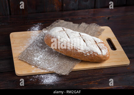 Frisches Brot auf einer Eiche board mit verstreuten Mehl auf einer hölzernen Hintergrund mit einem Stück Leinen Stoff. Stockfoto