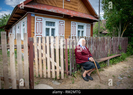Ältere Frau mit Kopftuch sitzt auf der Bank mit den Händen über Zuckerrohr vor der Kabine gekreuzt - Stil mit dekorativen Rollläden in der 'Trześcianka Stockfoto