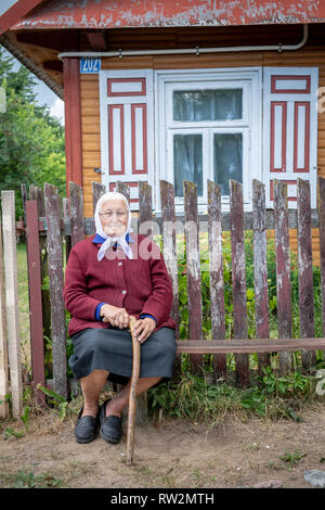 Ältere Frau mit Kopftuch sitzt auf der Werkbank mit Holz vor der Hütte - Stil mit dekorativen Fensterläden in Trześcianka das "Land der offenen Sh Stockfoto