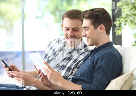 Glückliche Männer Kontrolle online Inhalte auf Tablet und Handy auf einer Couch im Wohnzimmer zu Hause sitzen Stockfoto