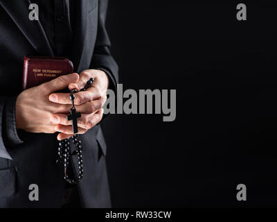 Die Hände eines christlichen Priesters in Schwarz mit einem Kruzifix und Neuen Testament Buch gekleidet. Religiöse Person mit Bibel und Gebet Perlen, Low-key-Bild wi Stockfoto