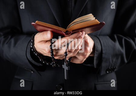 Die Hände eines christlichen Priesters in Schwarz mit einem Kruzifix und lesen Neue Testament Buch gekleidet. Religiöse person Studien Bibel und Gebet Perlen, Stockfoto