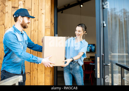 Delivery Man, die verpackte Ware mit einer jungen Frau Client gemeinsam im Freien vor der modernen Haus. Stockfoto