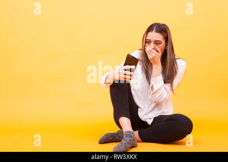 Mädchen suchen auf Ihr Telefon während Sie schockiert über Gelb Hintergrund sitzen. Gesichtsausdruck Stockfoto