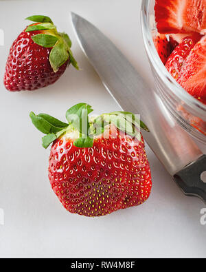 Nahaufnahme von Erdbeeren und ein Messer auf Weiß Schneidebrett. Teilweise Schüssel bereits in Scheiben geschnitten Beeren in der rechten oberen Ecke. Stockfoto